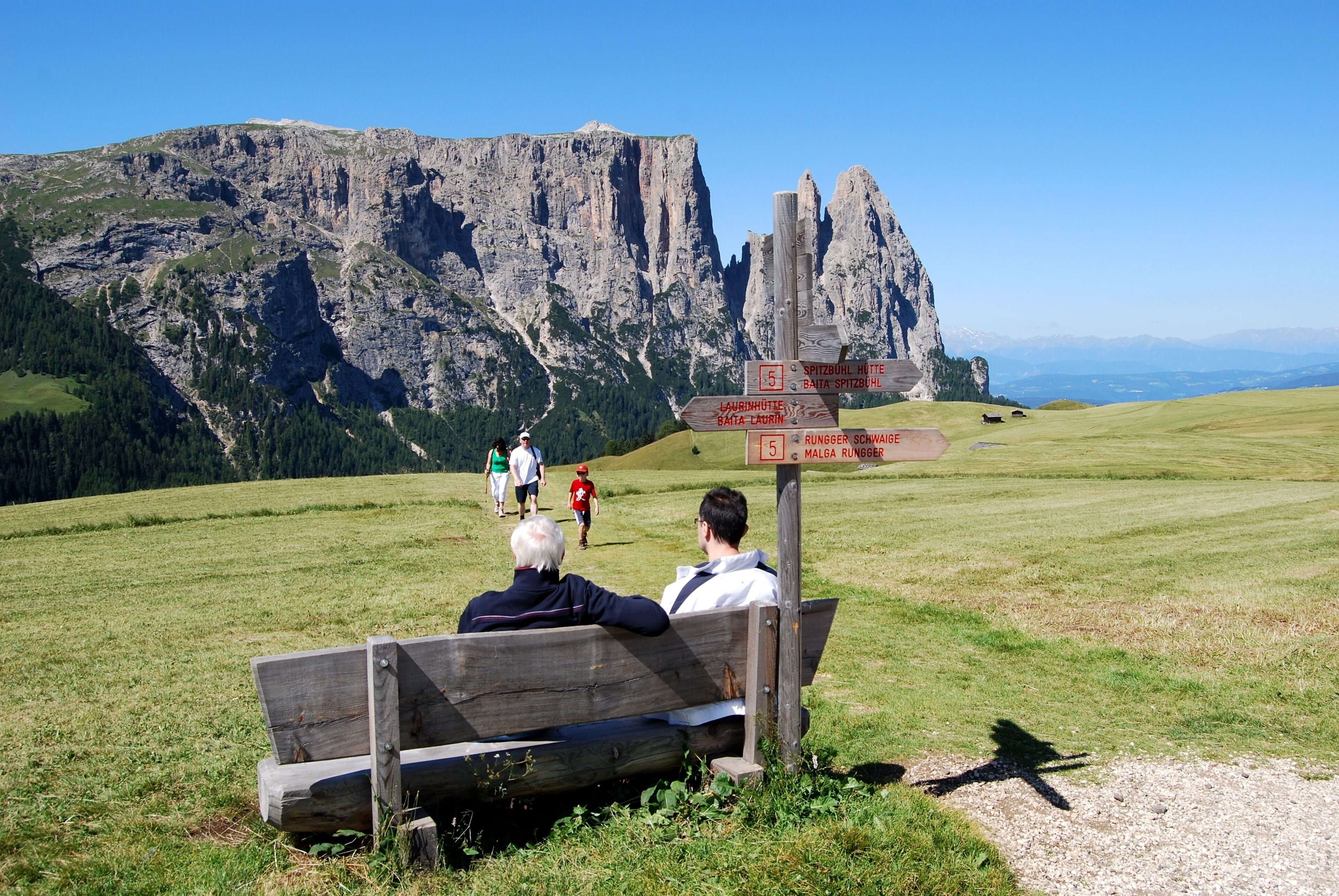 A wooden bench in a grassy field with a mountain backdrop and directional signs.