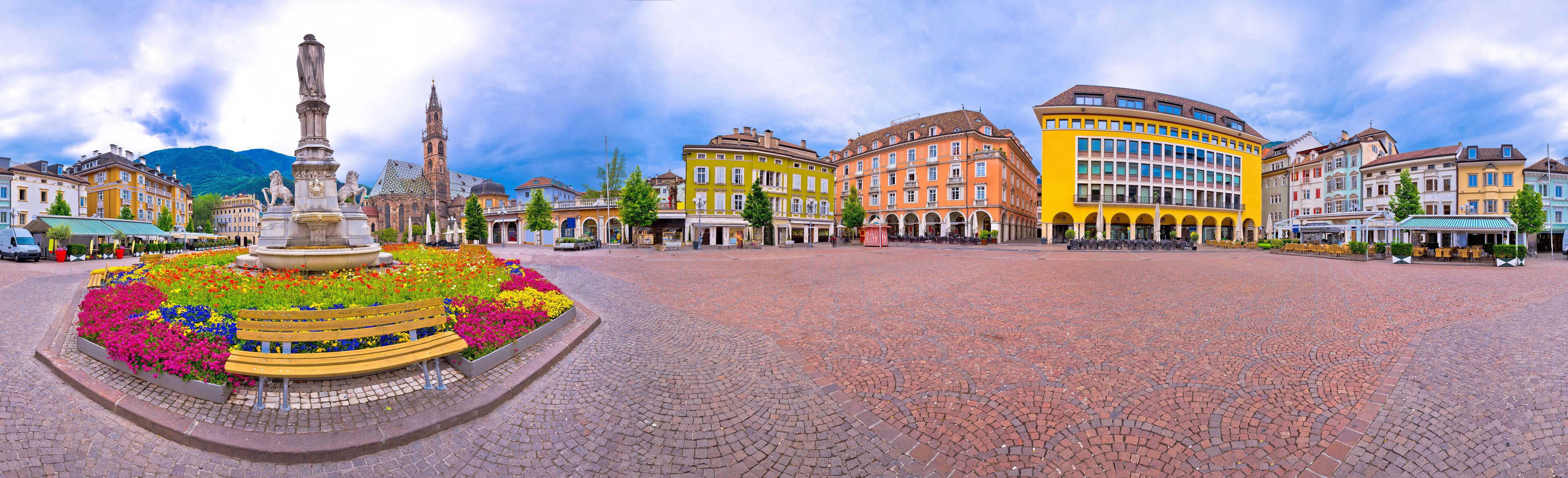 A cobblestone plaza with colorful flower beds, a statue, and historic buildings.