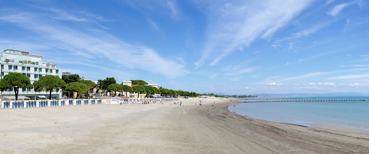 A sandy beach with a clear blue sky, a calm sea, and a row of buildings along the shore.