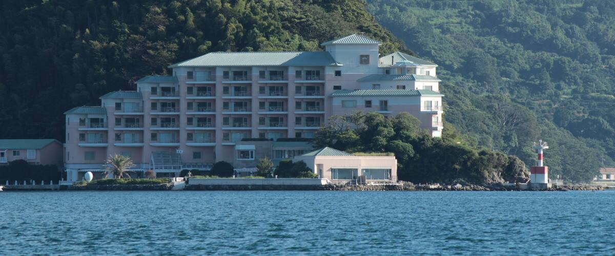 A coastal hotel with a green roof, surrounded by lush greenery and a lighthouse in the background.