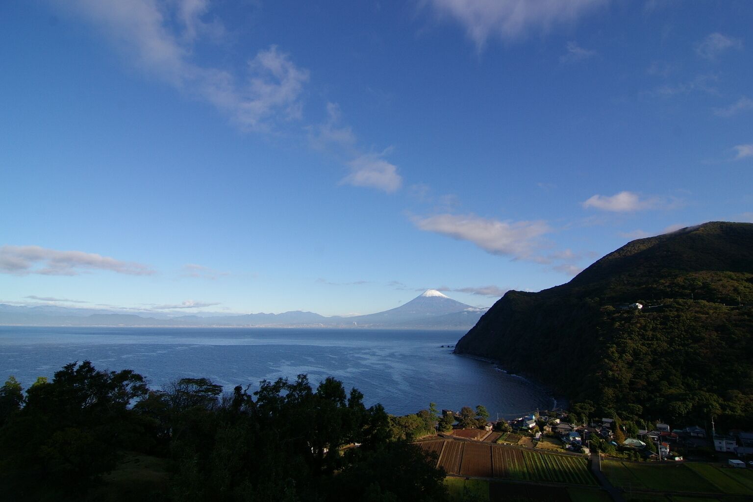 A coastal landscape with a mountain in the background, a body of water, and a village with houses and fields.