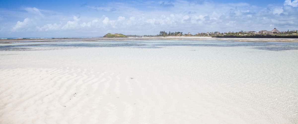 A sandy beach with clear water, a distant shoreline with buildings, and a partly cloudy sky.