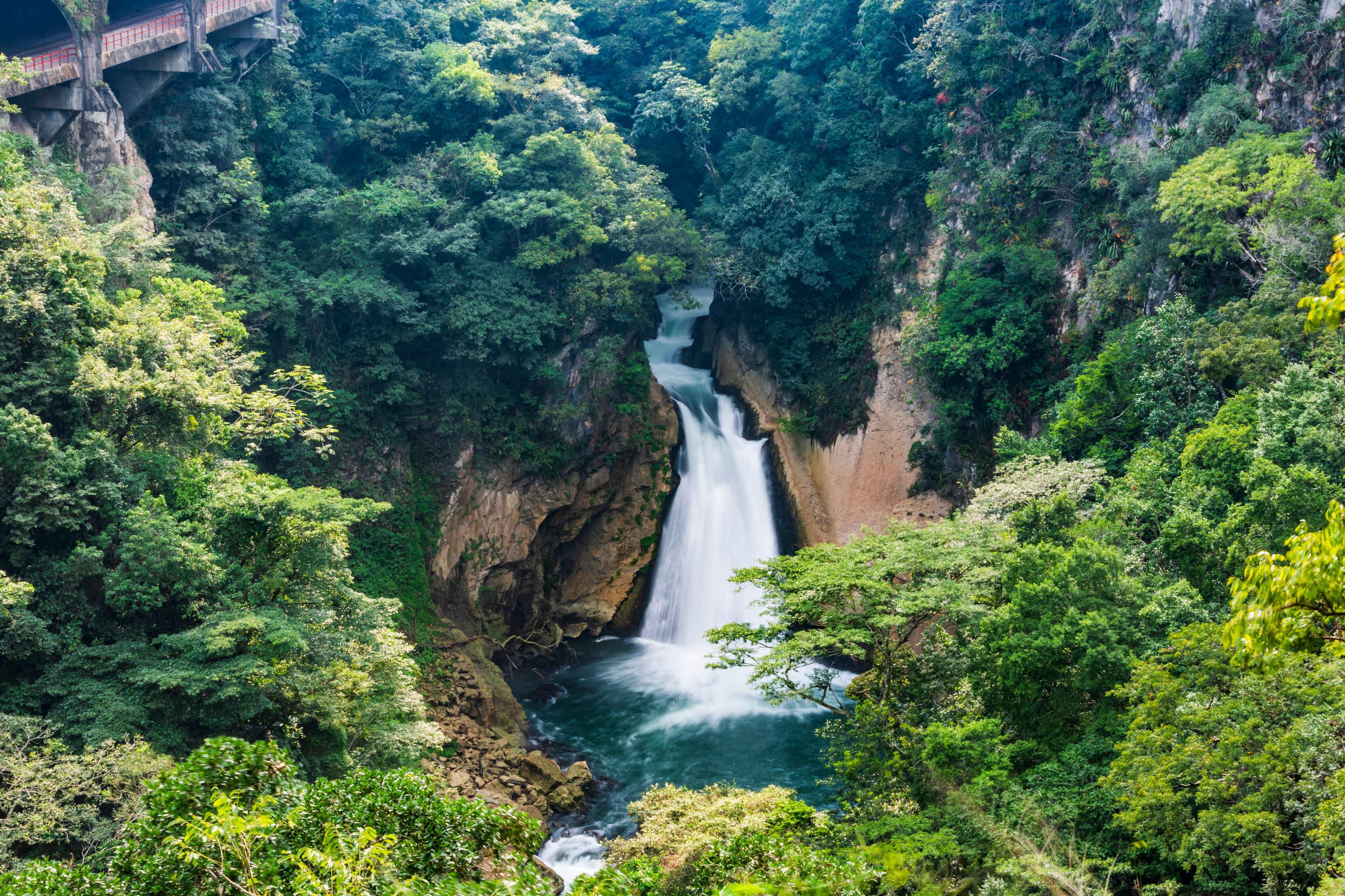 A waterfall in a lush forest with a bridge in the background.