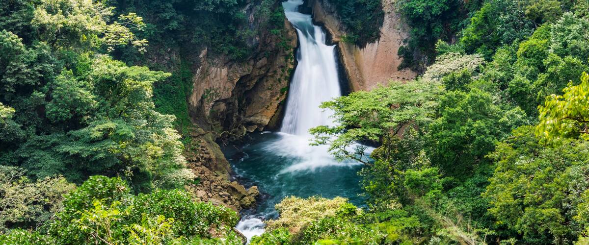A waterfall in a lush forest with a bridge in the background.