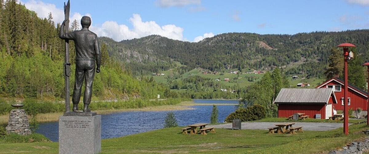 A statue of a man holding a rifle, a lake, and a red building with picnic tables.