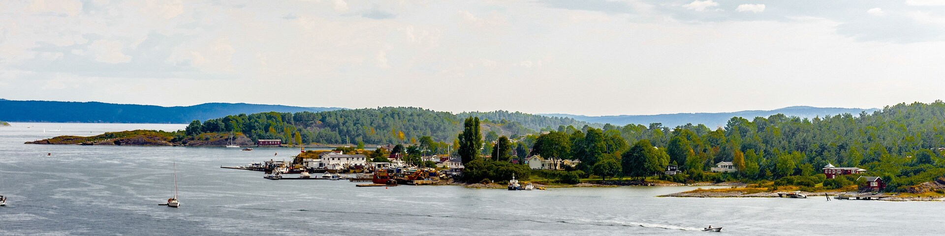 A coastal town with boats, a lighthouse, and a forested hill in the background.