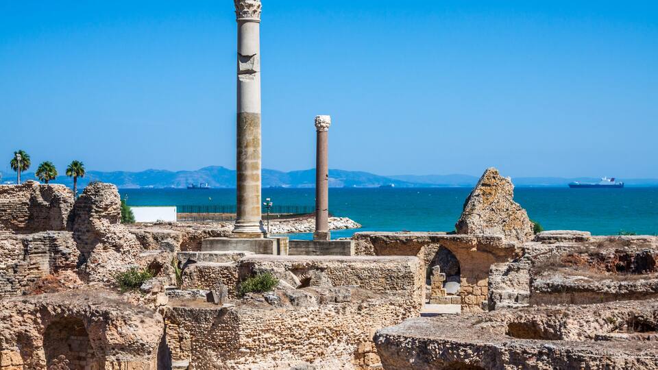 Ancient ruins with columns and arches, a clear blue sky, and the sea in the background.