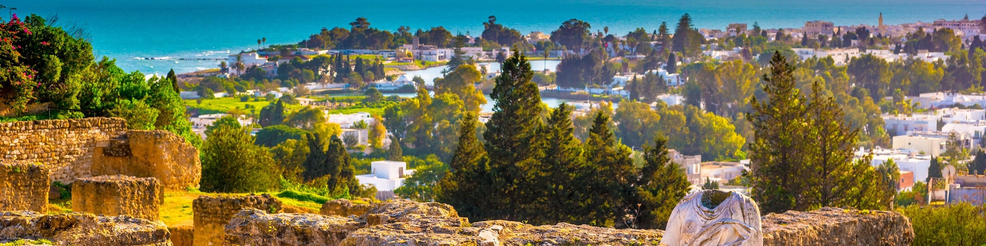 Ruínas antigas, uma vila costeira e uma montanha ao fundo.