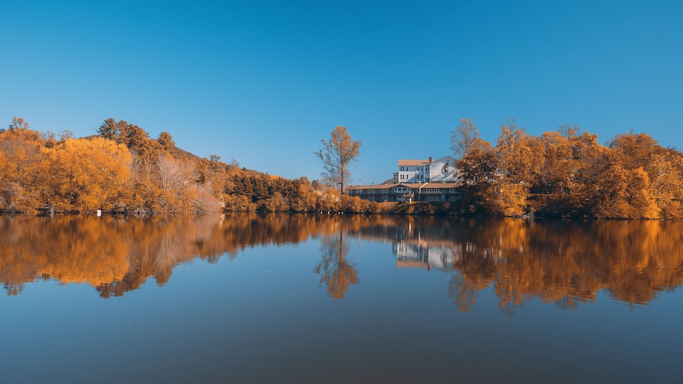 A calm lake with autumn-colored trees reflecting in the water, surrounded by a house and dense forest.
