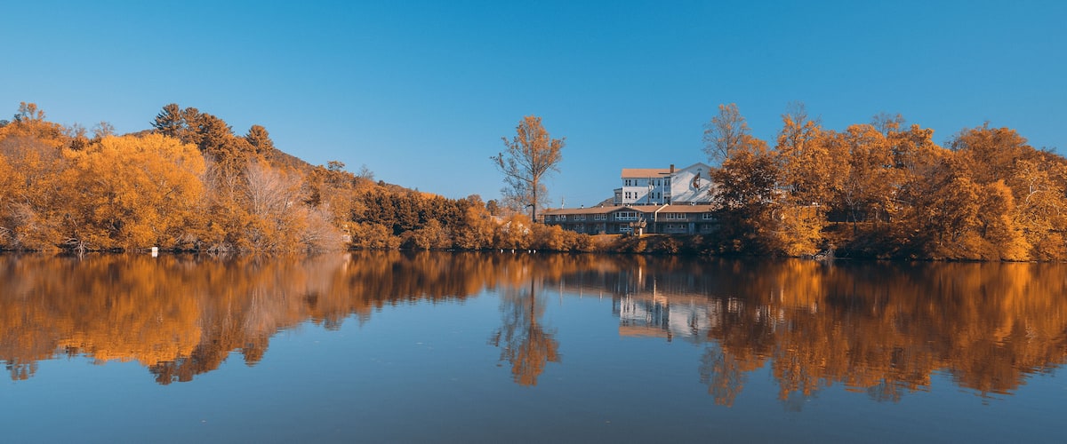 A calm lake with autumn-colored trees reflecting in the water, surrounded by a house and dense forest.
