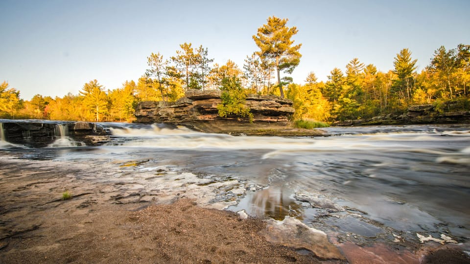 A river flowing over rocks with a waterfall, surrounded by autumn-colored trees.