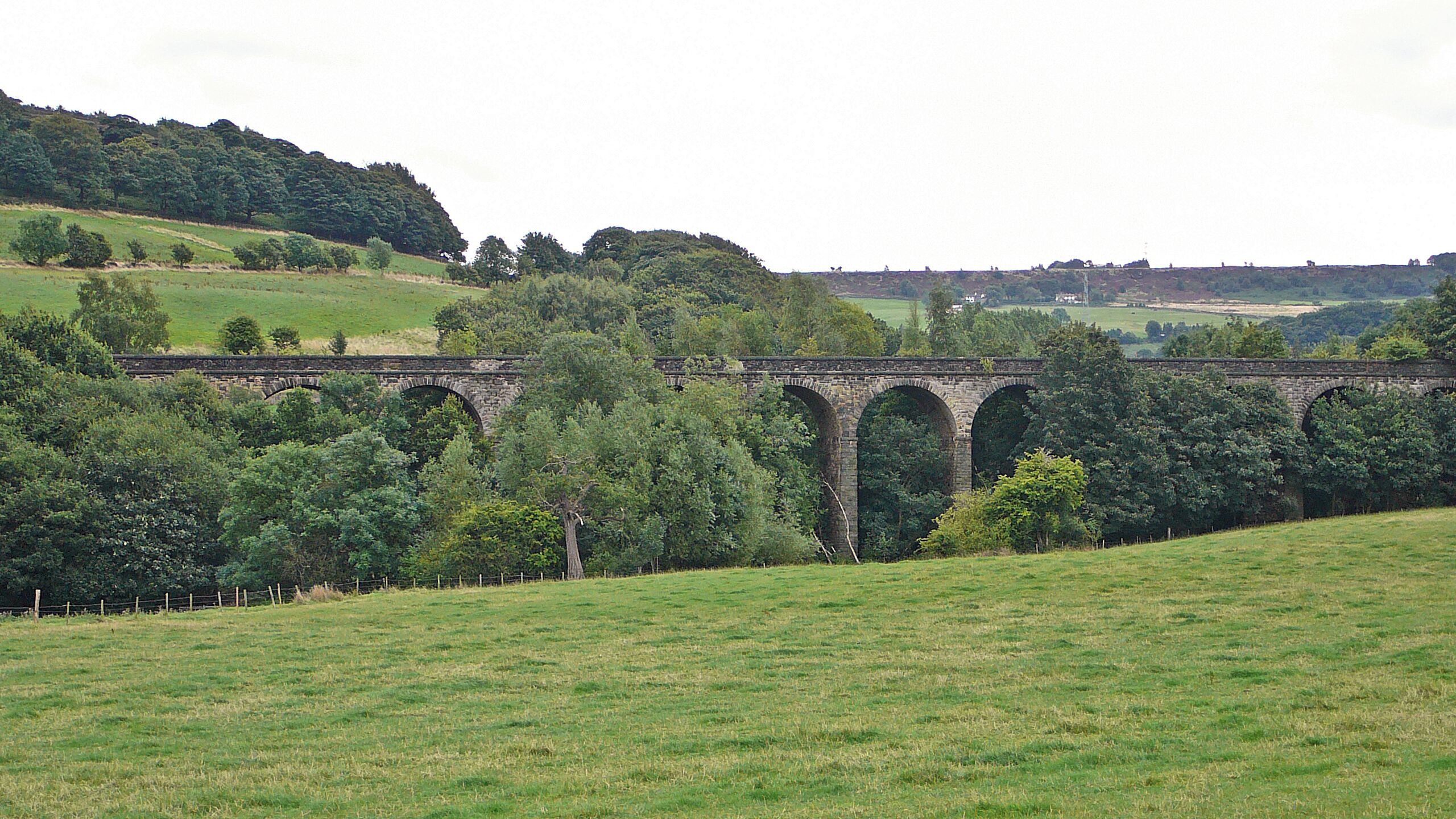 A stone viaduct over a grassy field with trees and hills in the background.