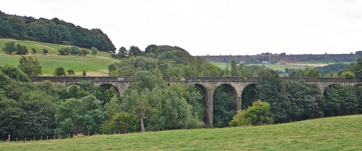 A stone viaduct over a grassy field with trees and hills in the background.