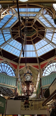 A large, ornate clock hanging from the ceiling of a market hall with a glass roof.