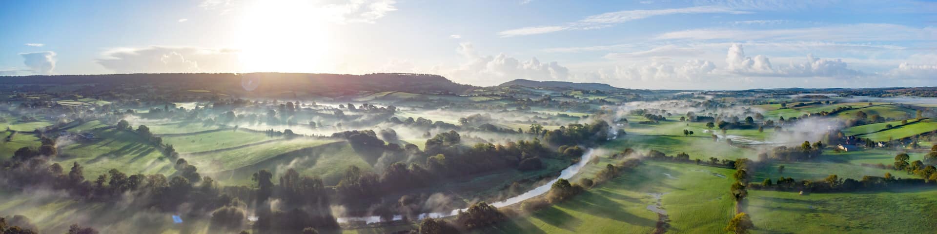 Aerial panorama picture of the river Otter near Honiton and Ottery St Mary. Sunrise and rolling mist cross the lush green fields below. Spectacular landscape.