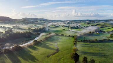 Aerial panorama picture of the river Otter near Honiton and Ottery St Mary. Sunrise and rolling mist cross the lush green fields below. Spectacular landscape.