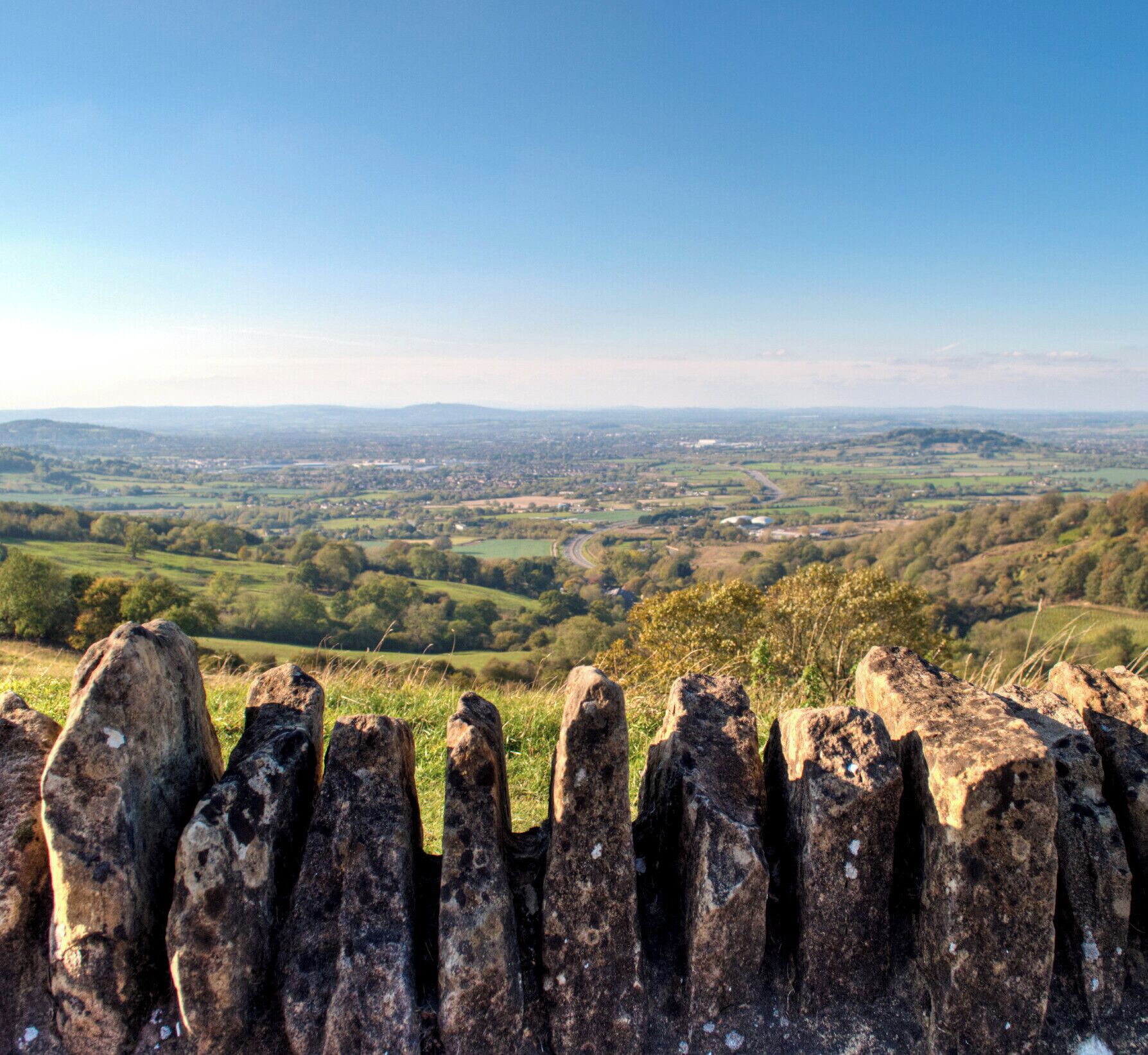 A rural landscape with rolling hills, a winding road, and a stone fence in the foreground.