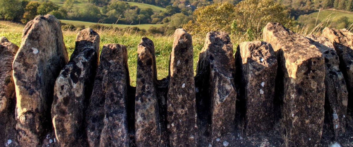A rural landscape with rolling hills, a winding road, and a stone fence in the foreground.