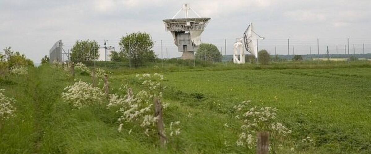 A large white satellite dish in a grassy field with wind turbines in the background.