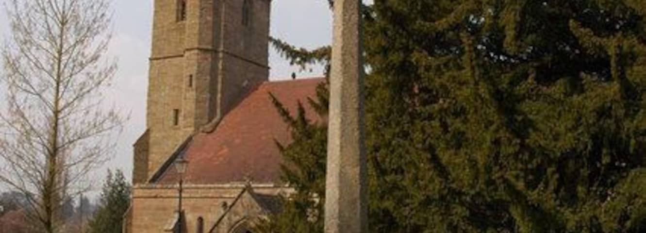 Stone cross in Holy Trinity parish churchyard, Belbroughton, Worcestershire, seen from the southeast, with the church steeple in the background. The cross is late Medieval, and was restored early in the 20th century as a monument to Major-General Sir Edward Woodgate, who was killed in 1900 in the Second South African War.