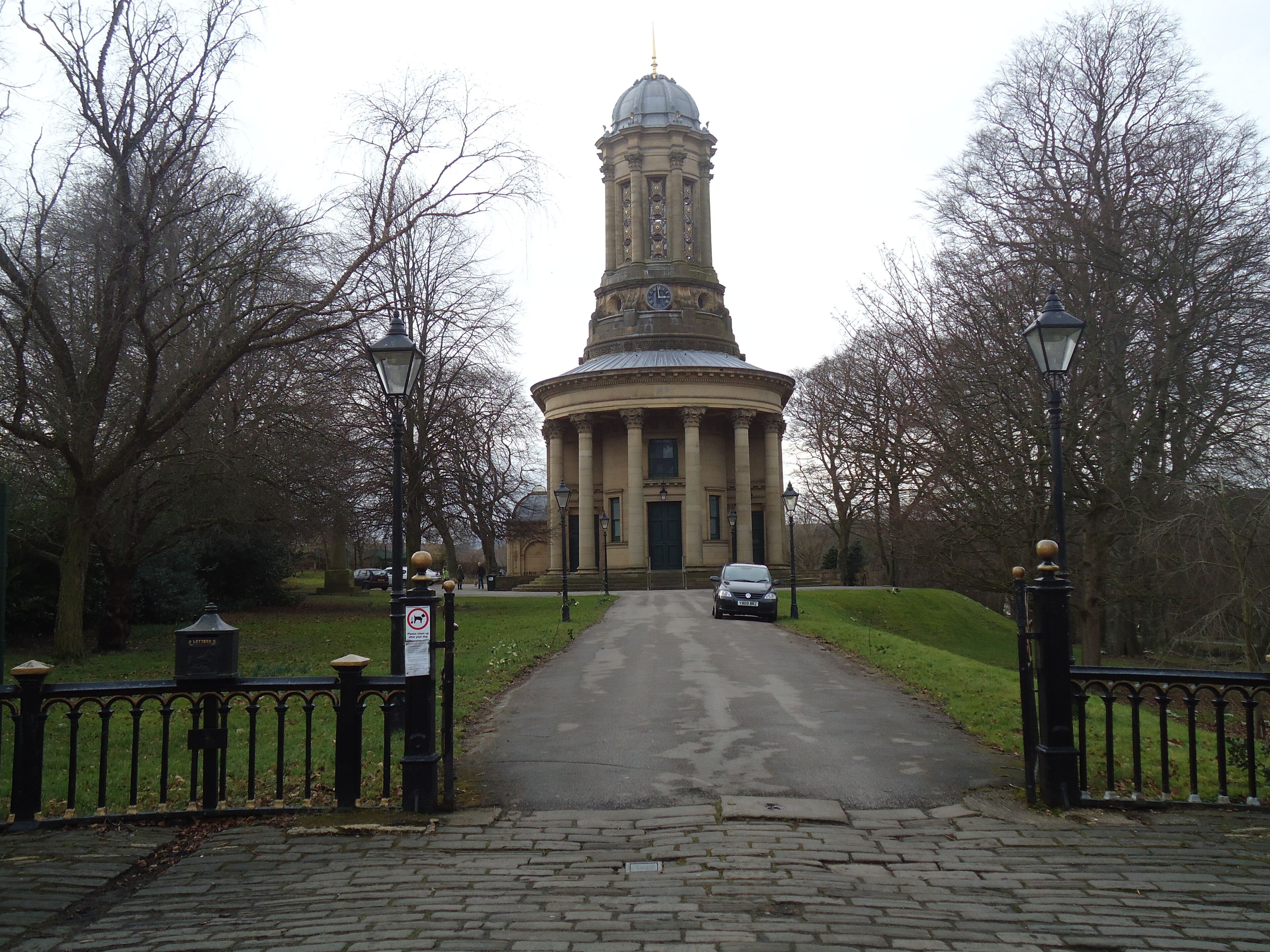 A historic building with a central dome and a clock tower, surrounded by a paved pathway and trees.