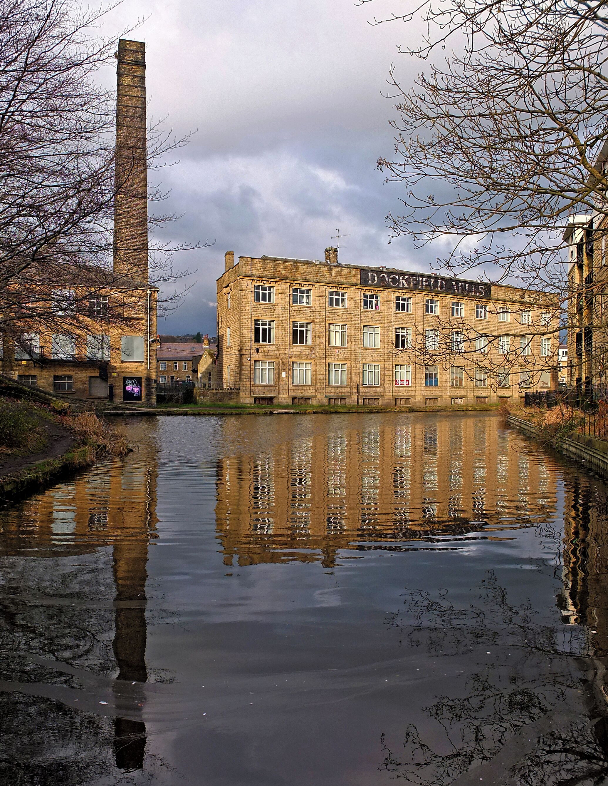 A historic building with a sign reading "Dockyard Mills" across the front, a tall chimney to the left, and a calm river reflecting the structure.