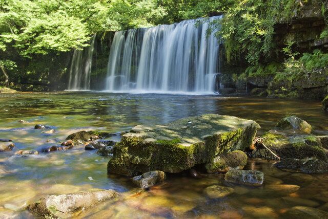 A waterfall cascading over rocks into a clear stream surrounded by lush greenery.