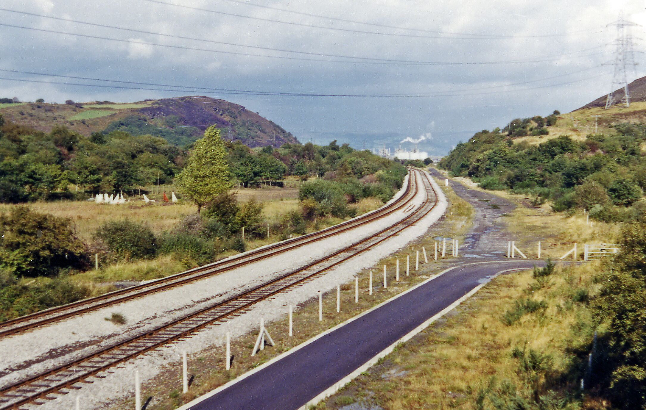 A railway track curving through a hilly landscape with greenery and a power line tower in the distance.