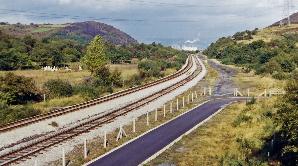 A railway track curving through a hilly landscape with greenery and a power line tower in the distance.