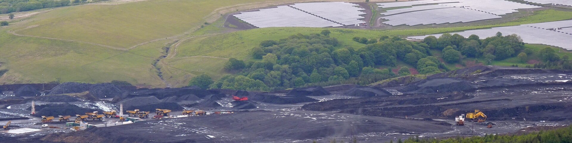 Operação de mineração com extração de carvão a céu aberto, cercada por colinas arborizadas e campos agrícolas.