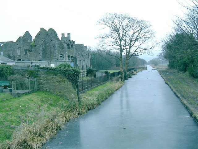 A narrow paved path flanked by grassy areas and a stone wall, leading to a historic stone building.
