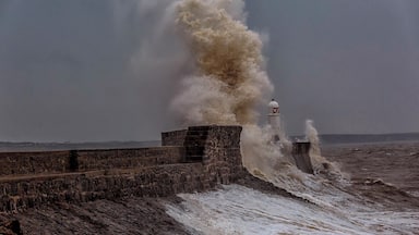 Une mer déchaînée s’écrase contre une jetée en pierre, près d’un phare.