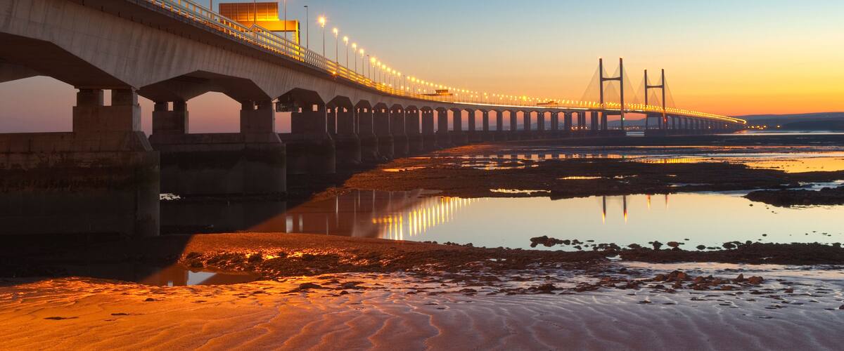 A long bridge with multiple arches and lights, spanning over a calm body of water at sunset.