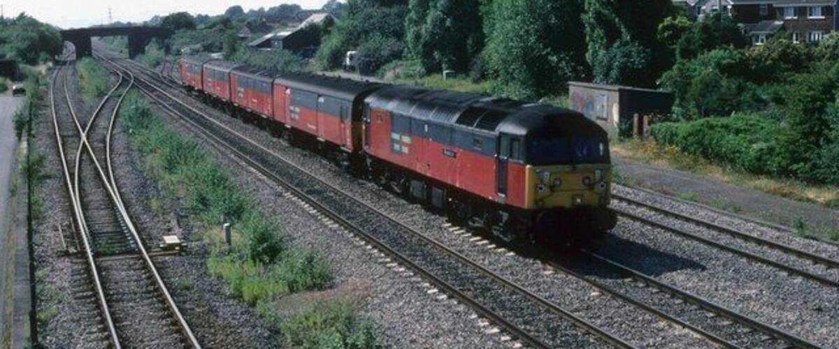 A red and black train on a railway track with houses and trees in the background.