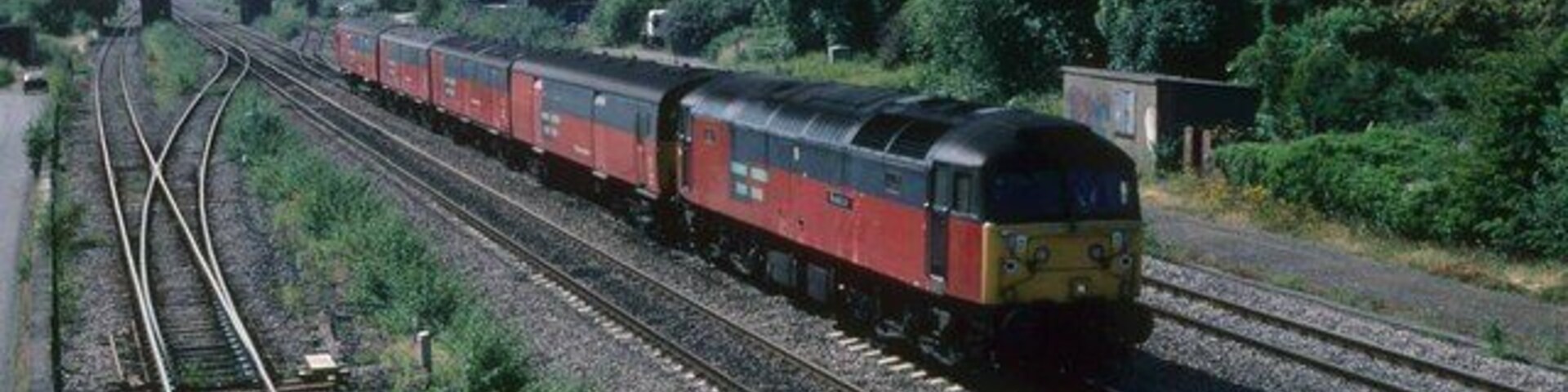 A red and black train on a railway track with houses and trees in the background.