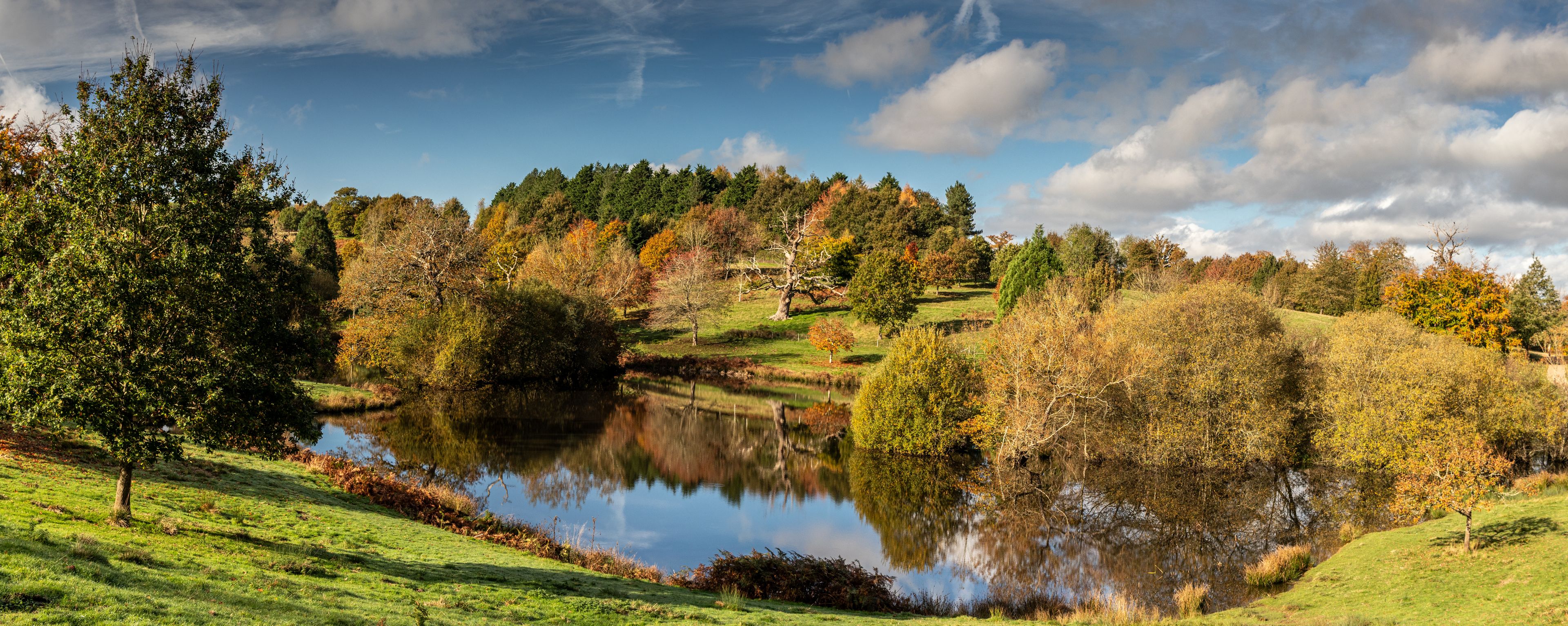 A serene lake surrounded by autumn foliage, a grassy foreground, and a hill with trees in the background.