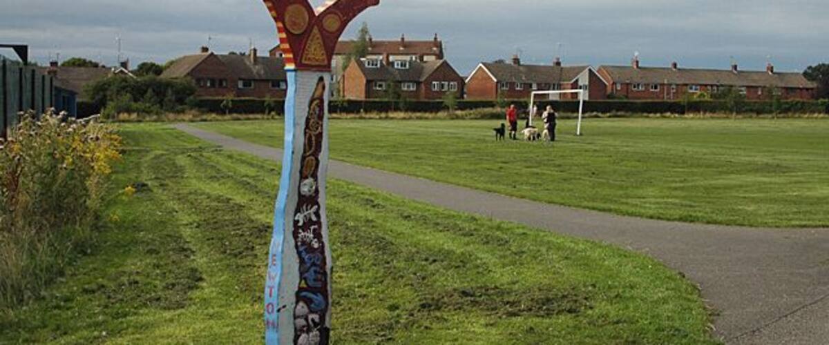 A grassy field with a path, houses in the background, and a tall, colorful sculpture.