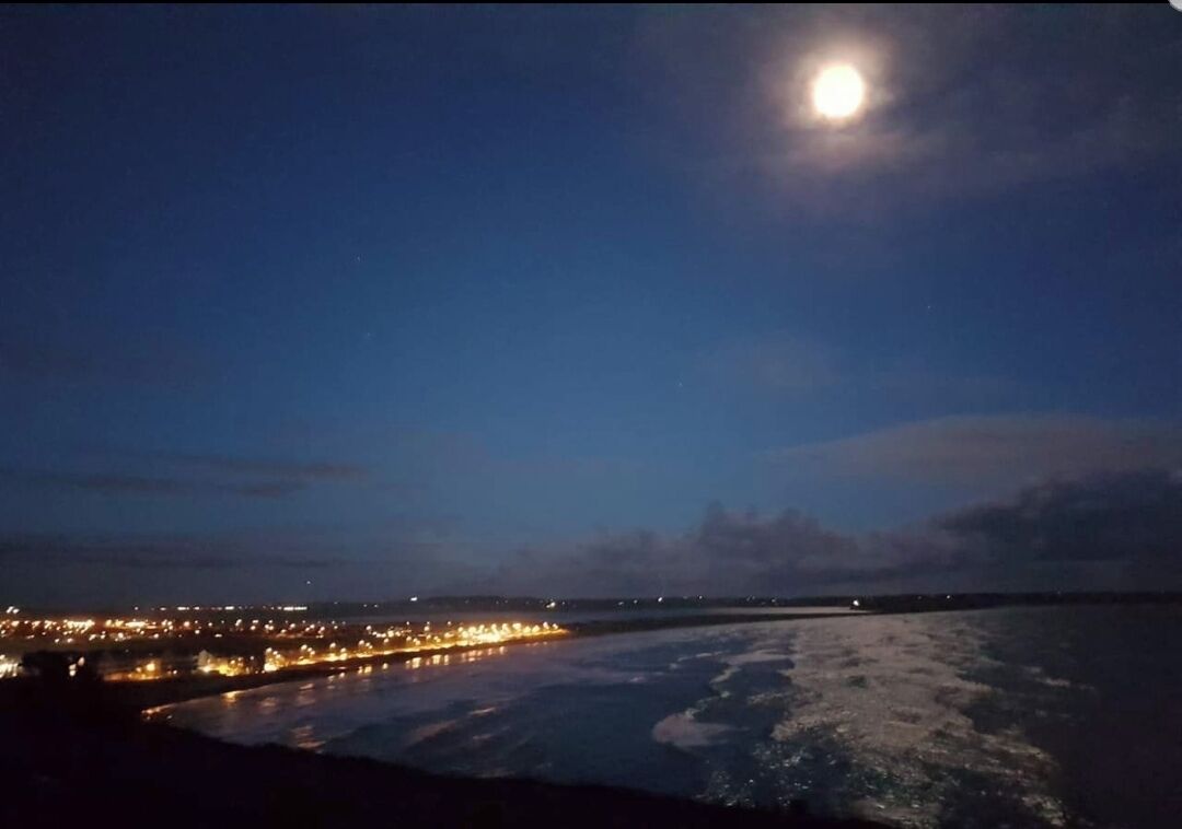 A night scene with a full moon, a coastline with city lights, and calm sea waves.