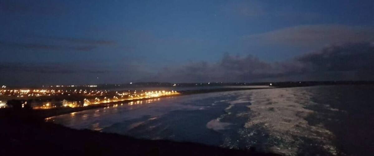 A night scene with a full moon, a coastline with city lights, and calm sea waves.