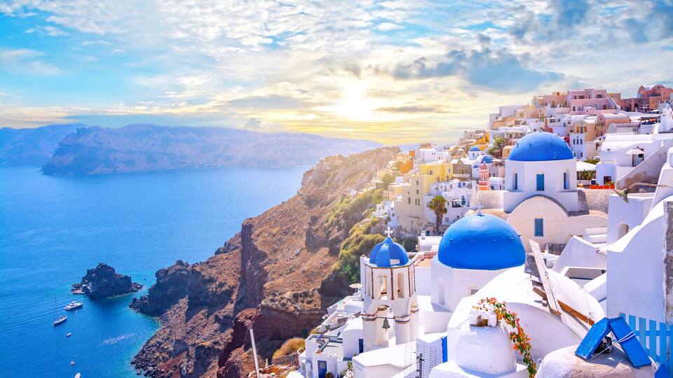 A coastal town with white buildings and blue domes, a rocky coastline, and boats in the water.