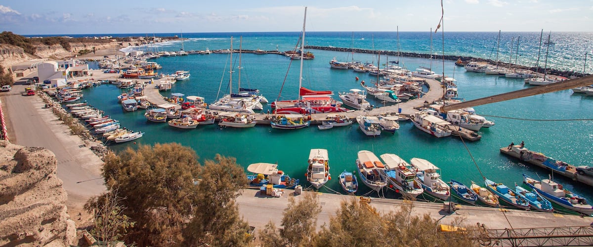 A marina with numerous boats docked in a circular arrangement, surrounded by a coastal landscape and a clear blue sky.