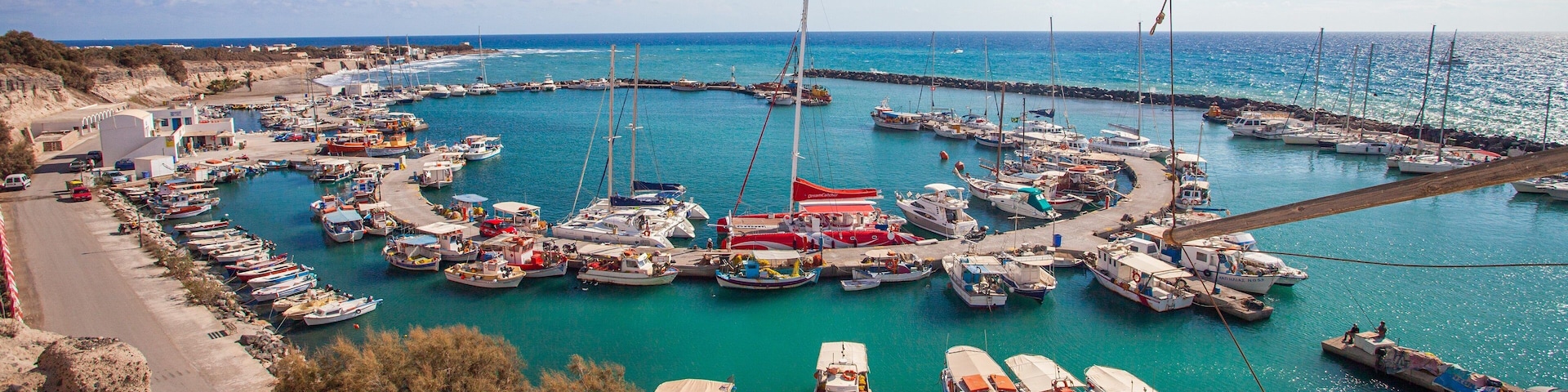 A marina with numerous boats docked in a circular arrangement, surrounded by a coastal landscape and a clear blue sky.