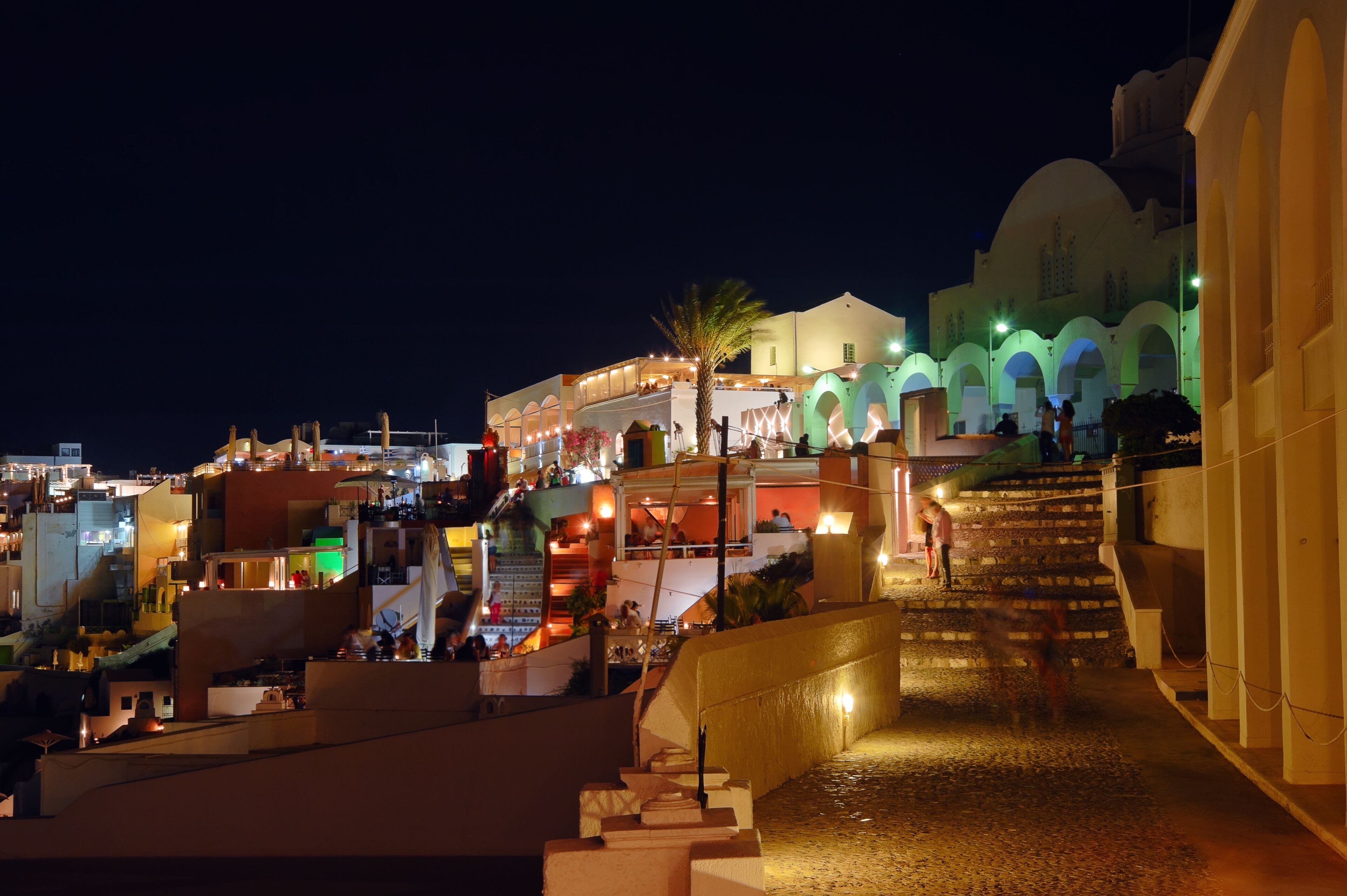 Una escena nocturna de un pueblo costero con edificios iluminados, una escalera que conduce a una mezquita y una palmera.