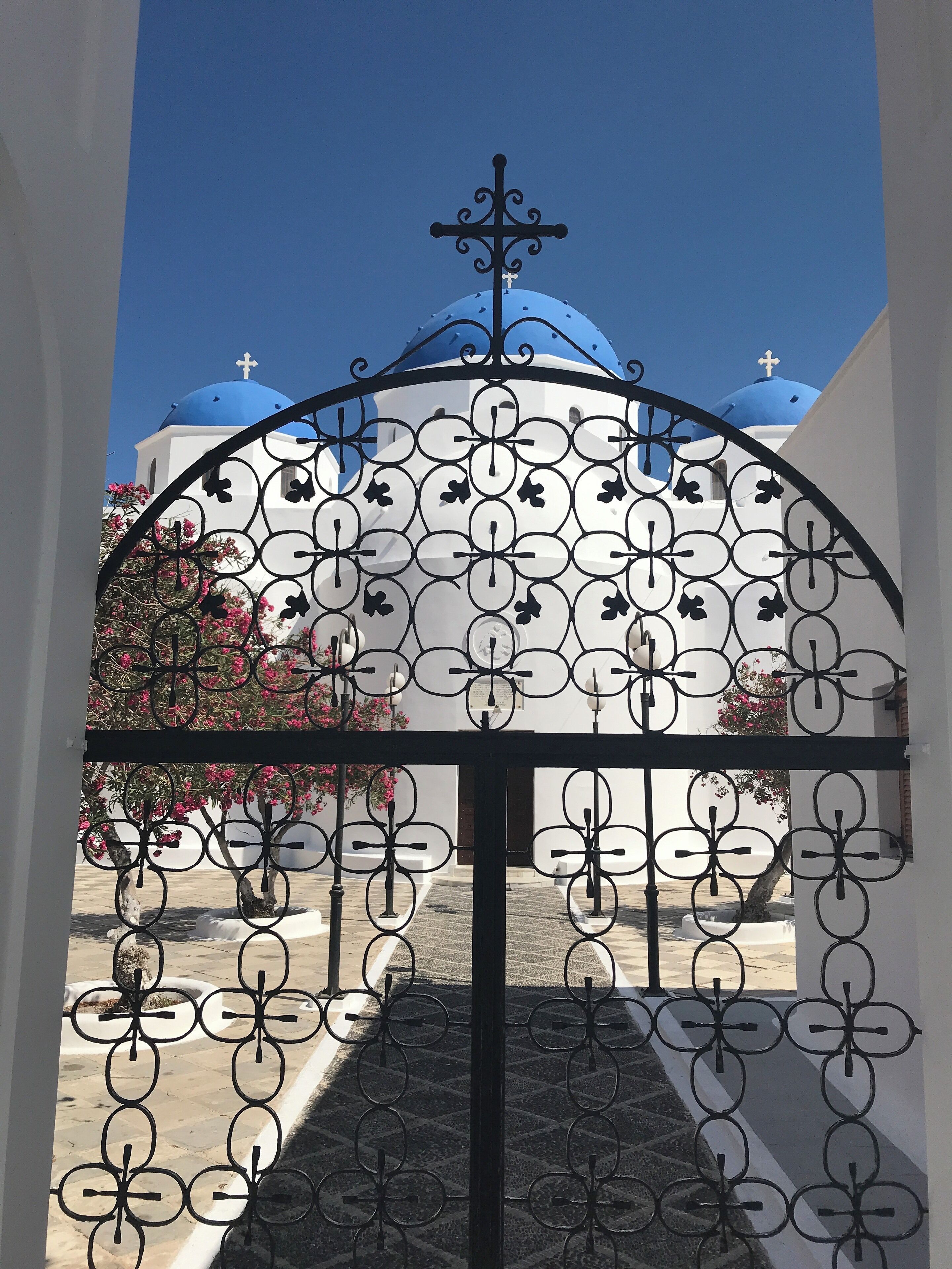 A wrought iron gate with a cross at the top, leading to a building with blue domes and crosses.