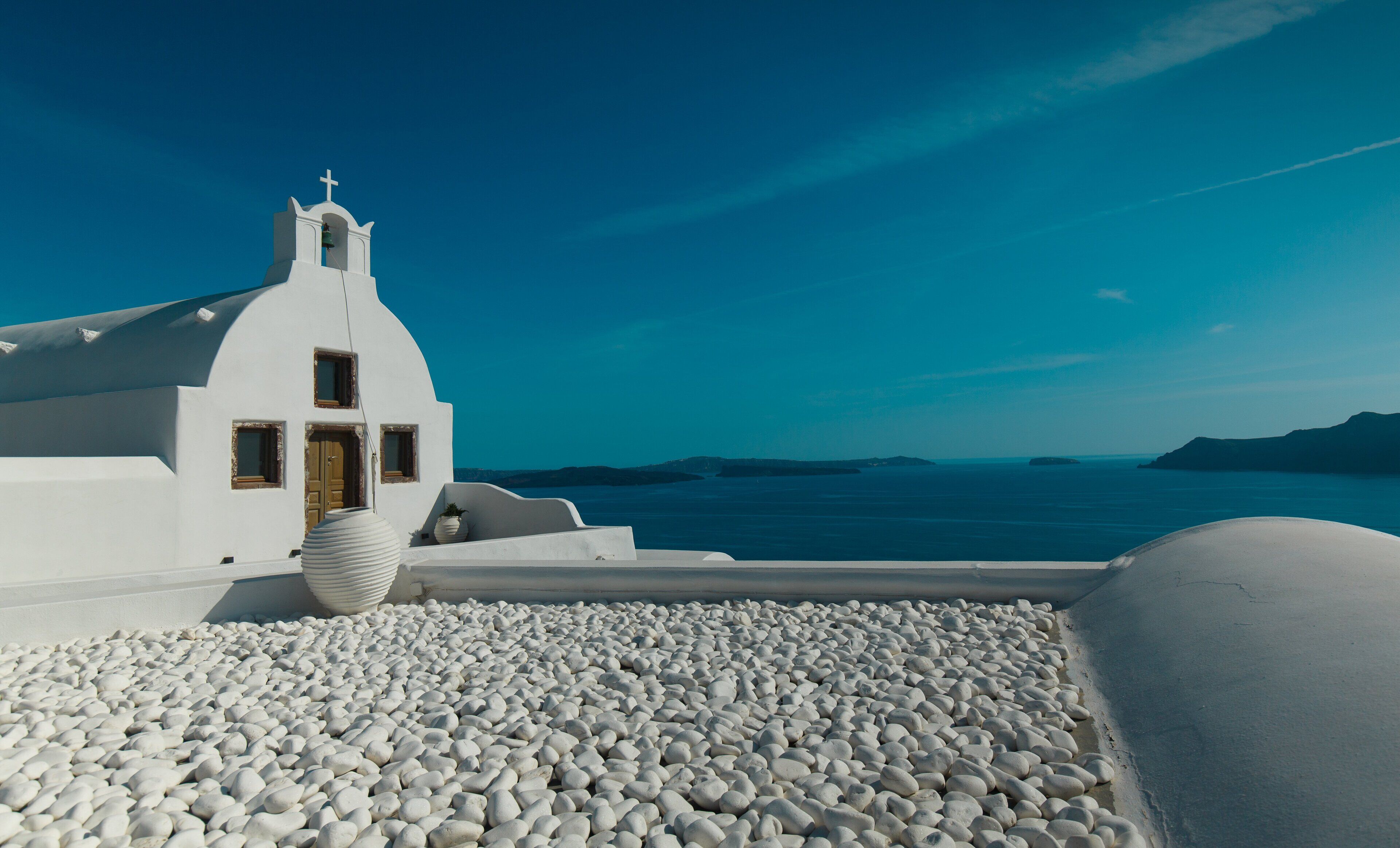 Un edificio blanco con un campanario y una puerta, rodeado de guijarros blancos y con vistas al mar azul.