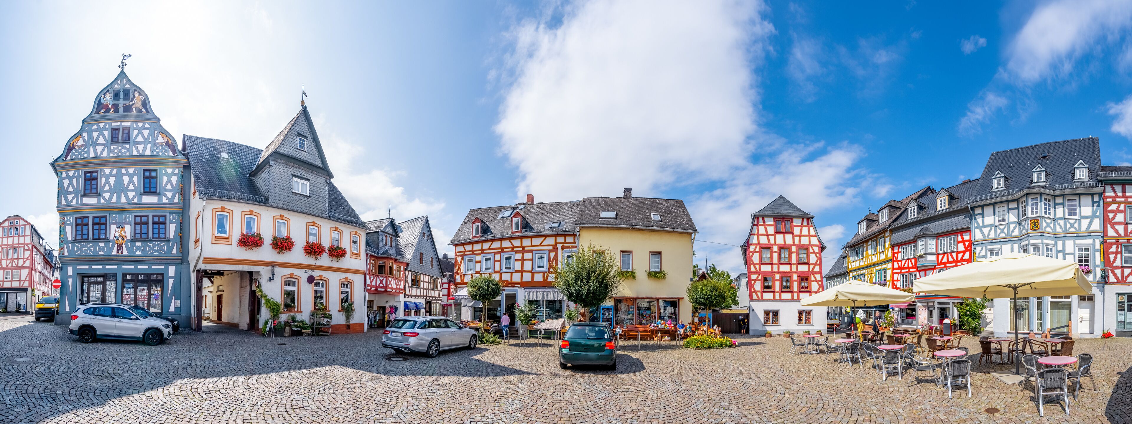 A cobblestone square with traditional half-timbered buildings, parked cars, and outdoor seating areas.
