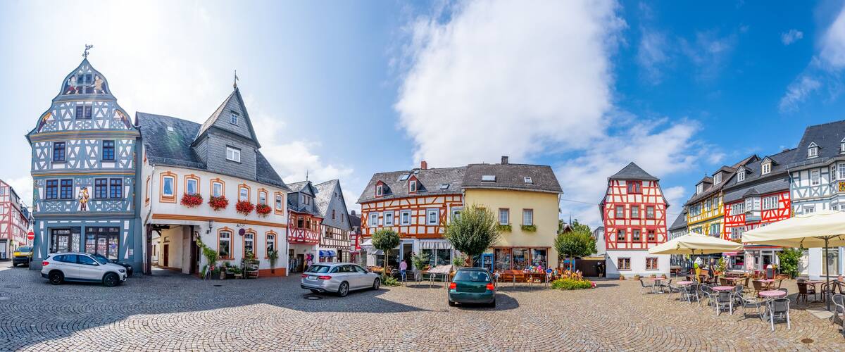 A cobblestone square with traditional half-timbered buildings, parked cars, and outdoor seating areas.