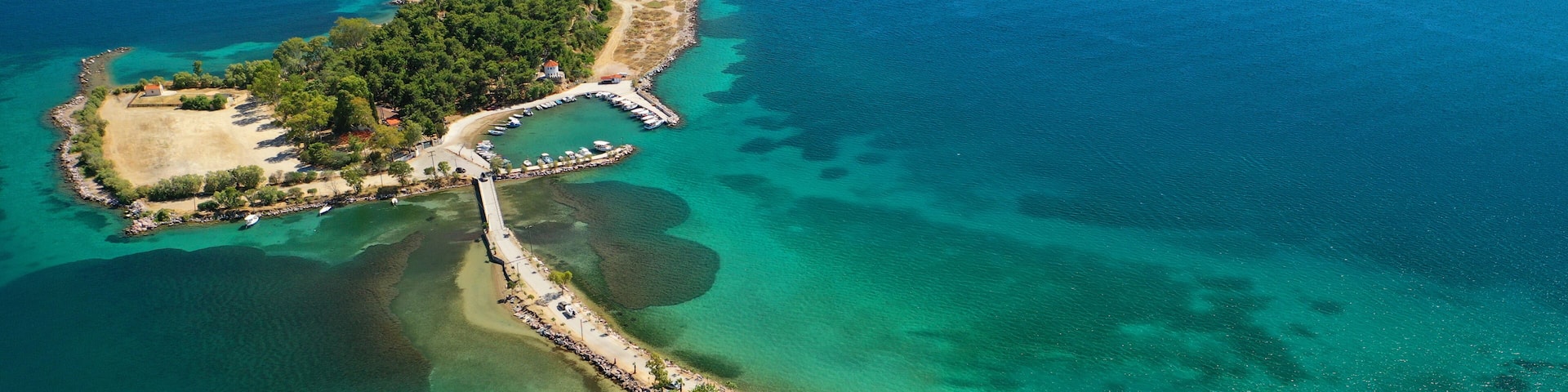 An aerial view of a coastal area with a pier, a small island with dense vegetation, clear turquoise waters, and a beach with umbrellas and cars.