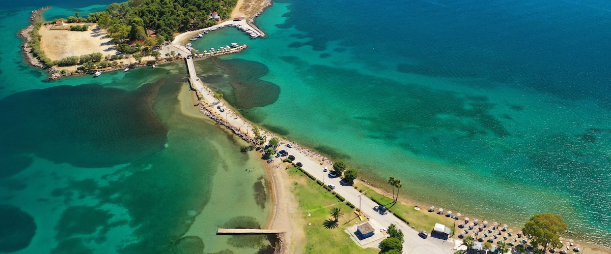 An aerial view of a coastal area with a pier, a small island with dense vegetation, clear turquoise waters, and a beach with umbrellas and cars.