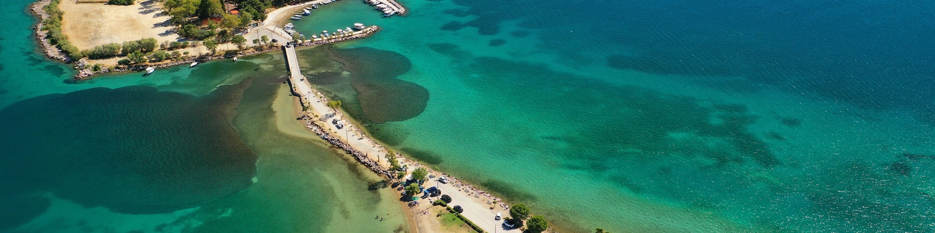 An aerial view of a coastal area with a pier, a small island with dense vegetation, clear turquoise waters, and a beach with umbrellas and cars.